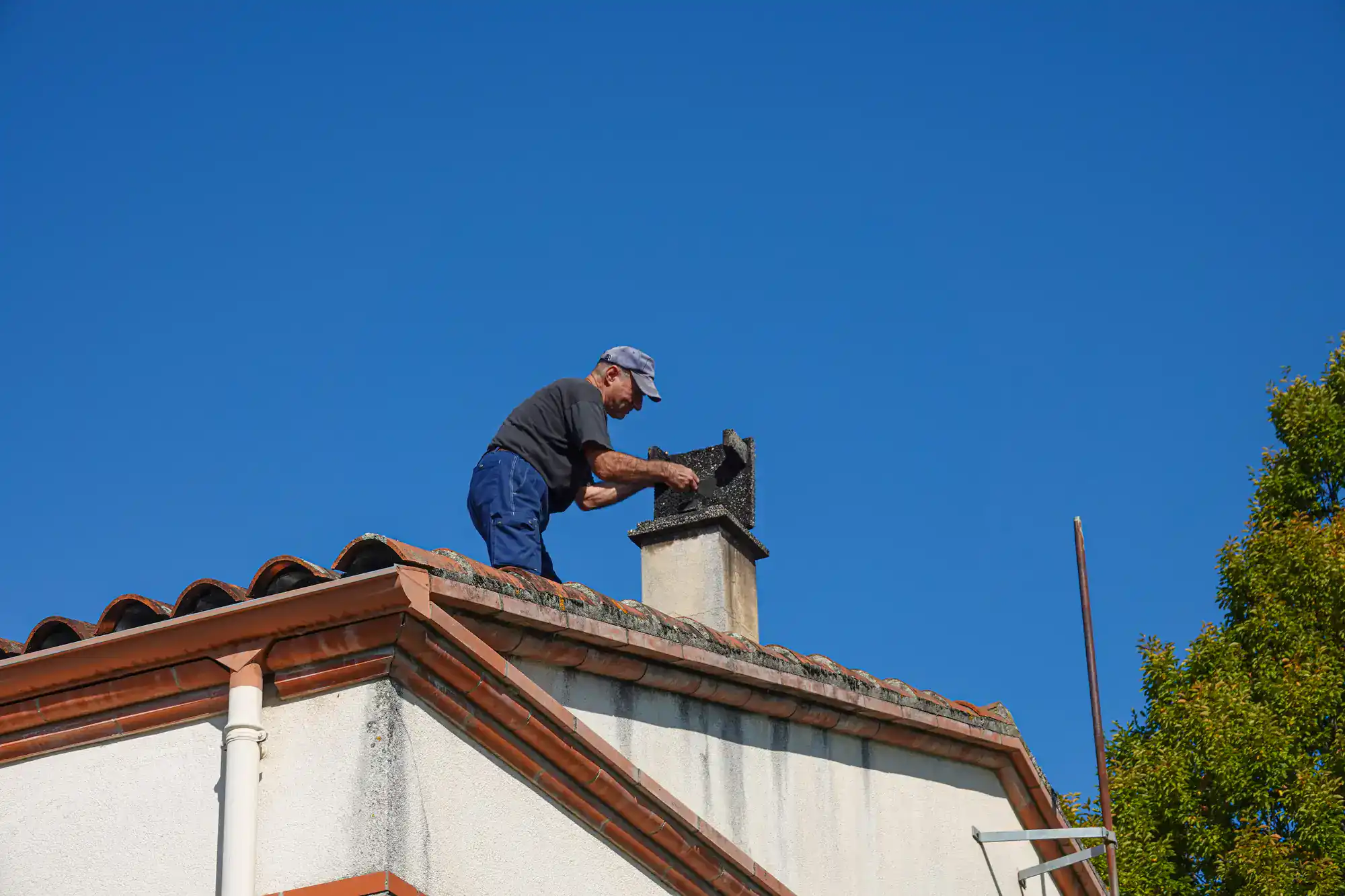 A man in a cap and blue pants is cleaning or repairing a chimney on the roof of a house under a clear blue sky. A ladder is positioned against the house and a tree is visible on the right side.