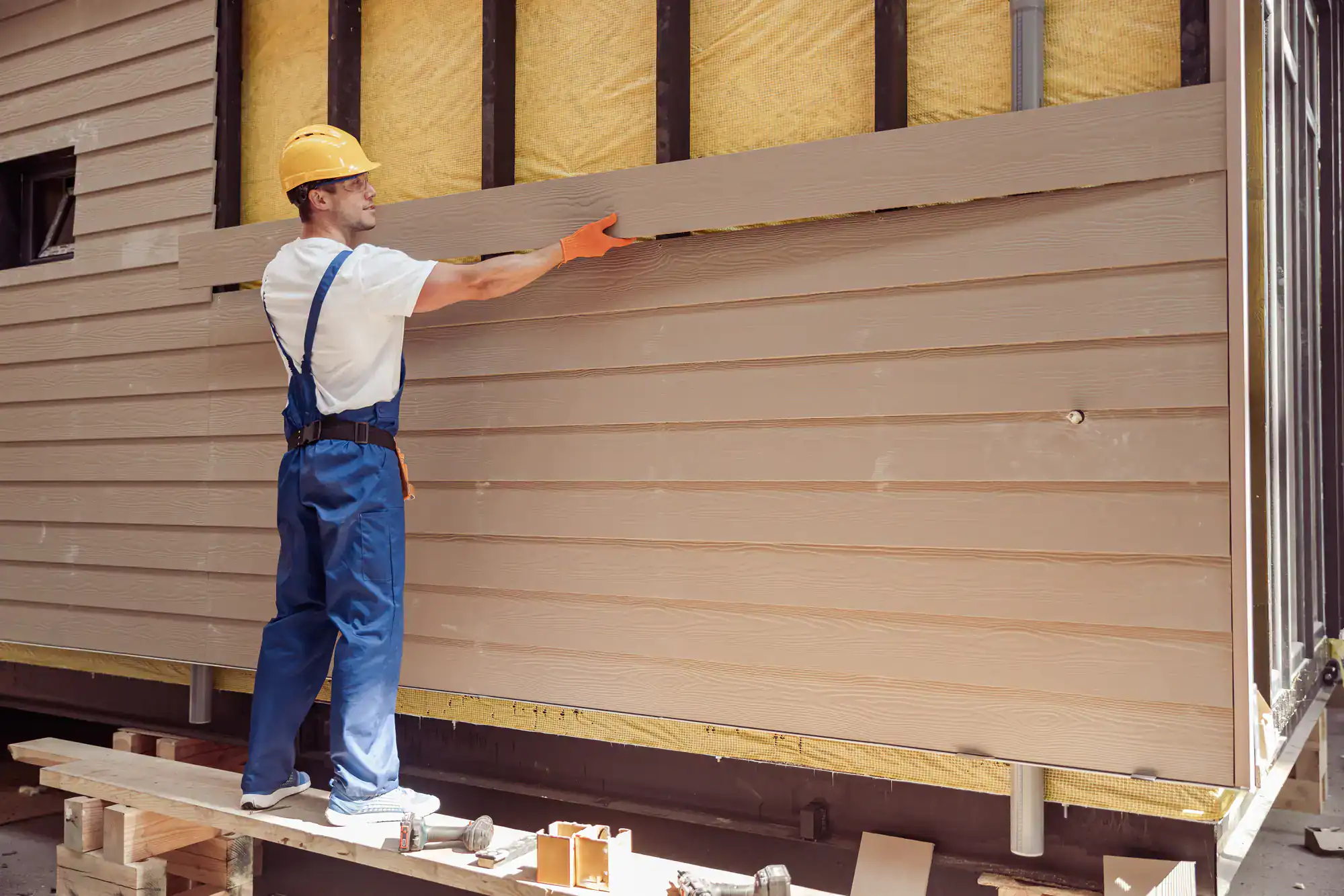 A construction worker in a yellow hard hat and blue overalls installs horizontal siding panels on a house exterior, with insulation and framing visible behind the new boards.