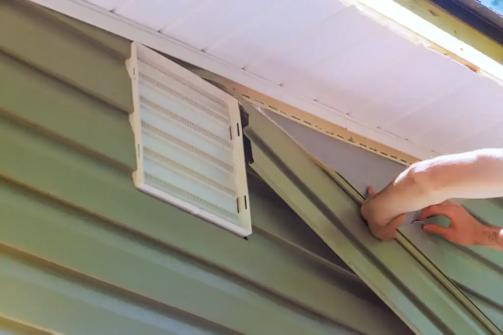 A person installs green vinyl siding on a house, aligning the panels under a white vent near the roof eaves.