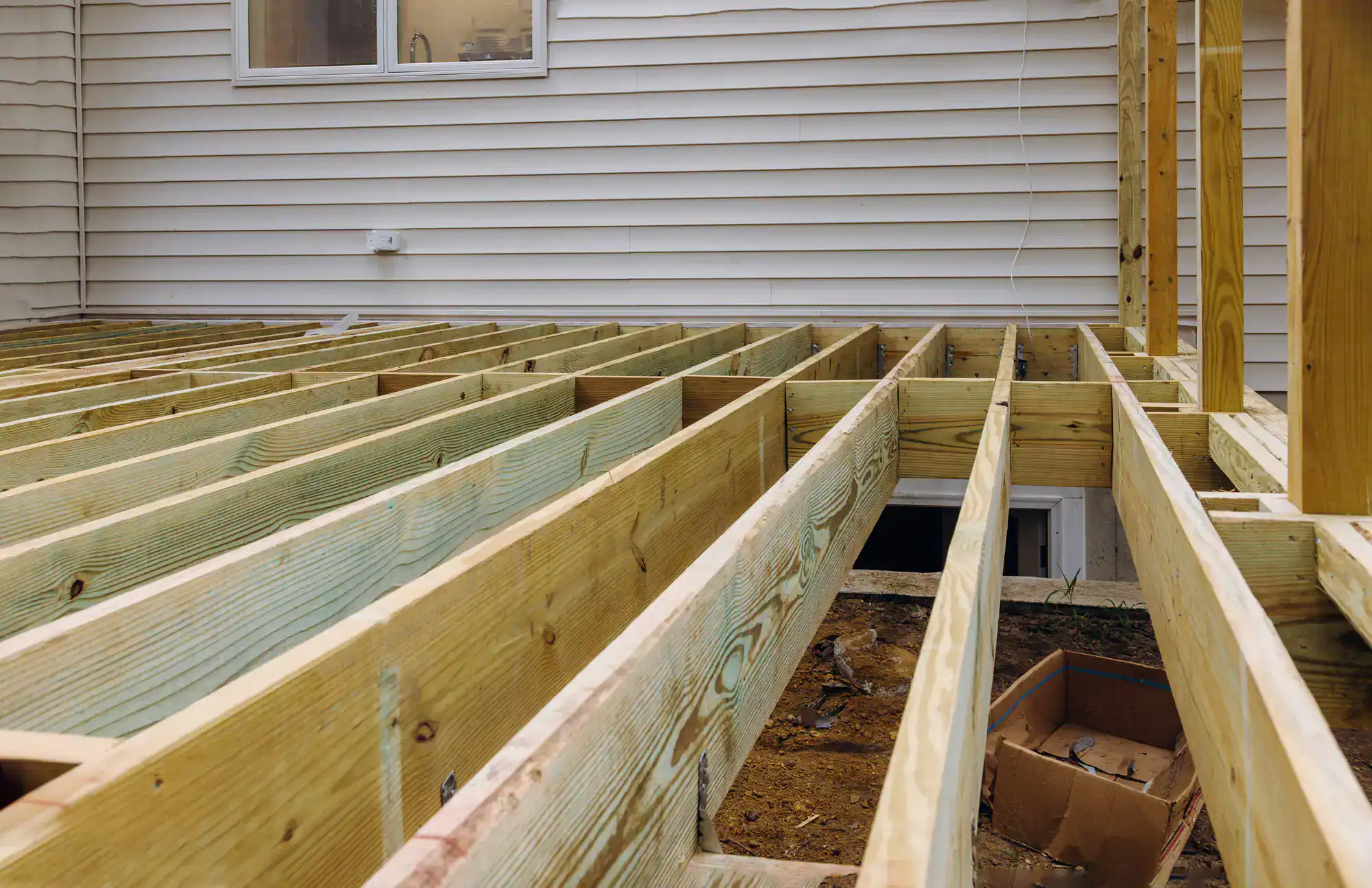 A wooden deck frame under construction is attached to a house with beige siding. Exposed beams and joists are visible, and a cardboard box is on the ground below the structure.
