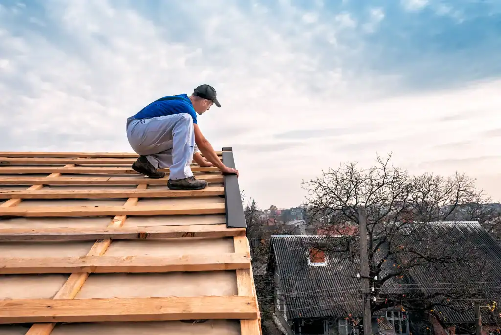 A person wearing a cap and work clothes installs roofing material on a wooden roof frame, with houses and leafless trees visible in the background under a partly cloudy sky.
